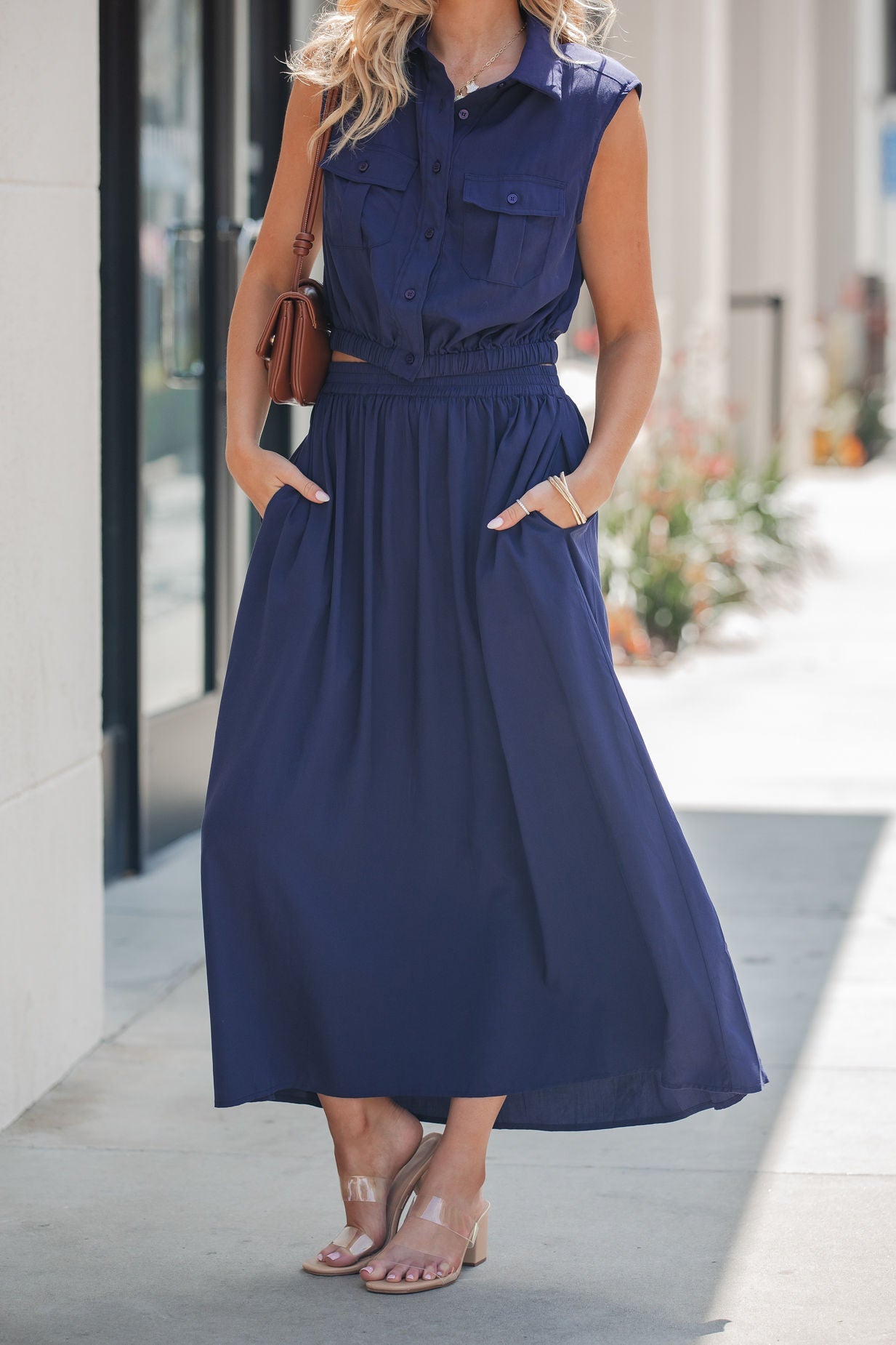 Woman in a sleeveless navy top and Elan Smocked Midi Skirt - Navy, holding a small brown purse on the sidewalk.