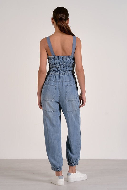 A woman faces away in the Elan Voyager Smocked Denim Jumpsuit, sleeveless and relaxed fit, styled with white sneakers.