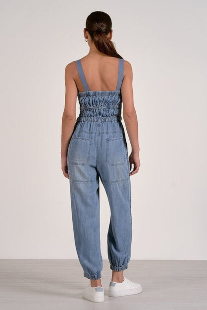A woman faces away in the Elan Voyager Smocked Denim Jumpsuit, sleeveless and relaxed fit, styled with white sneakers.
