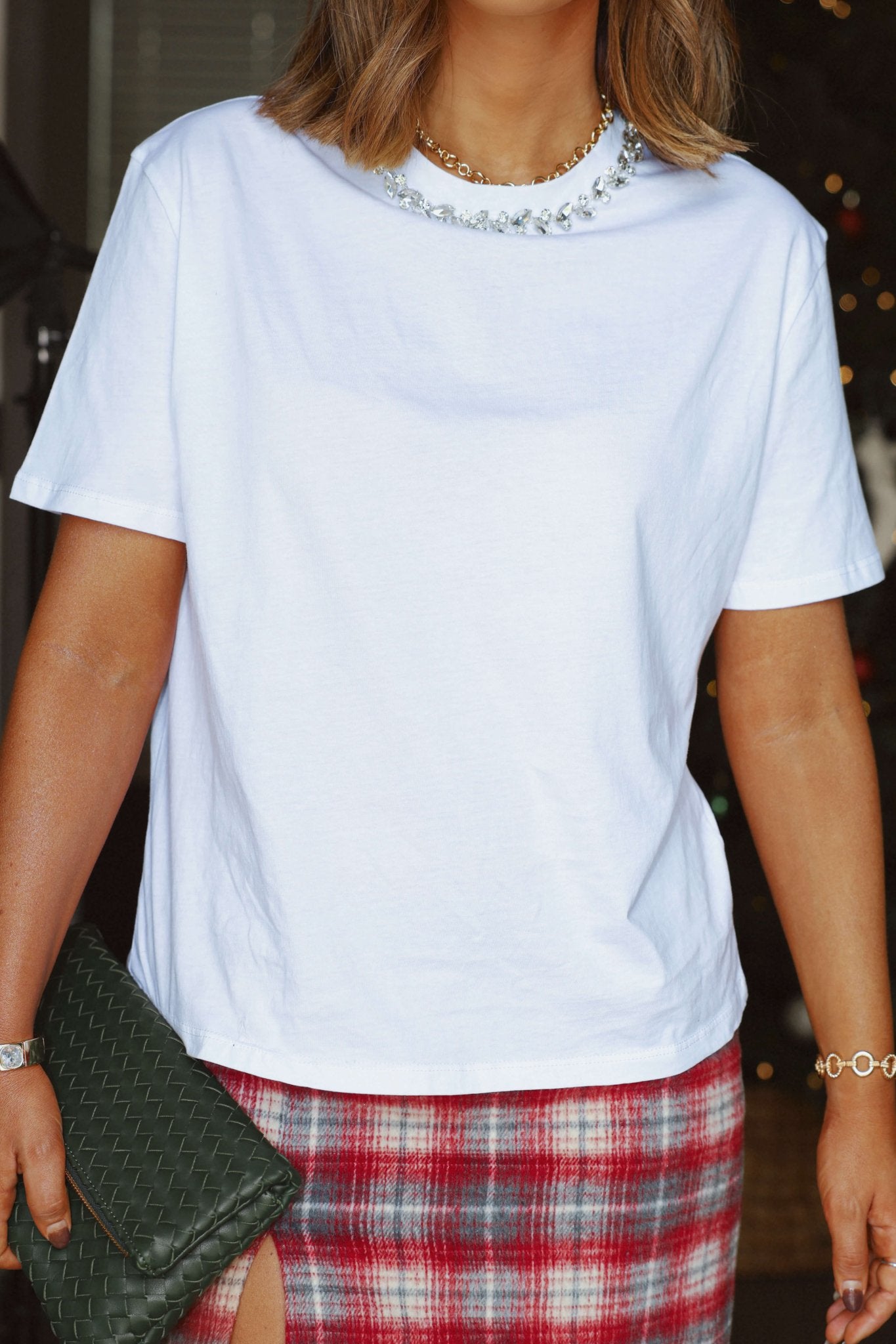 Woman wearing the Elan White Embellished Tee, a plaid red skirt, gold necklace, and holding a green textured clutch.