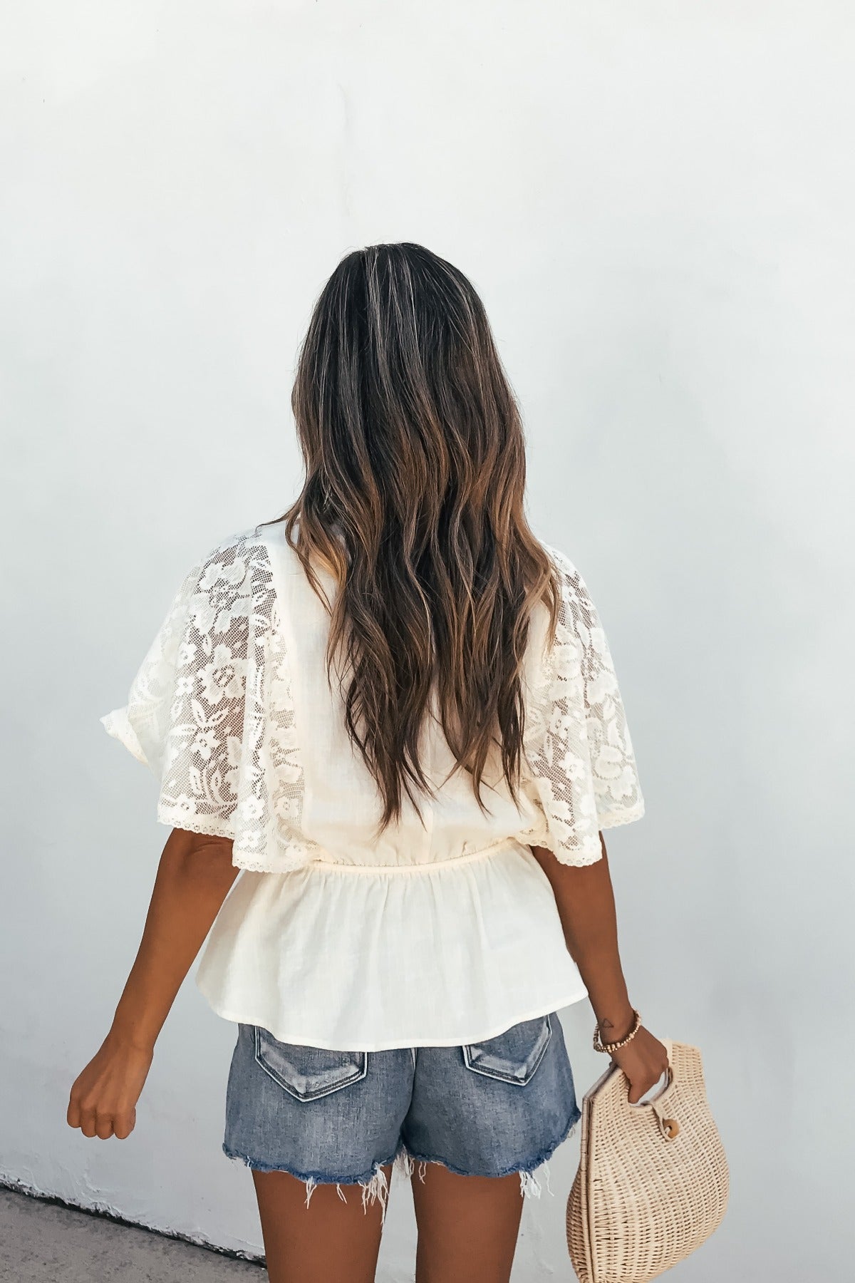 Woman with wavy brown hair in the Elanor Lace Peplum Top - Cream, denim shorts, and woven bag, facing a white wall.
