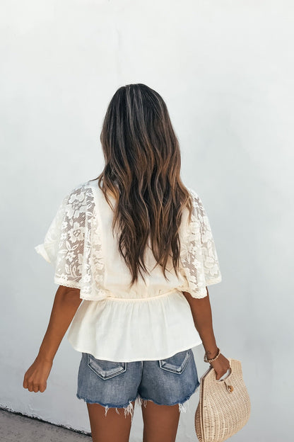Woman with wavy brown hair in the Elanor Lace Peplum Top - Cream, denim shorts, and woven bag, facing a white wall.