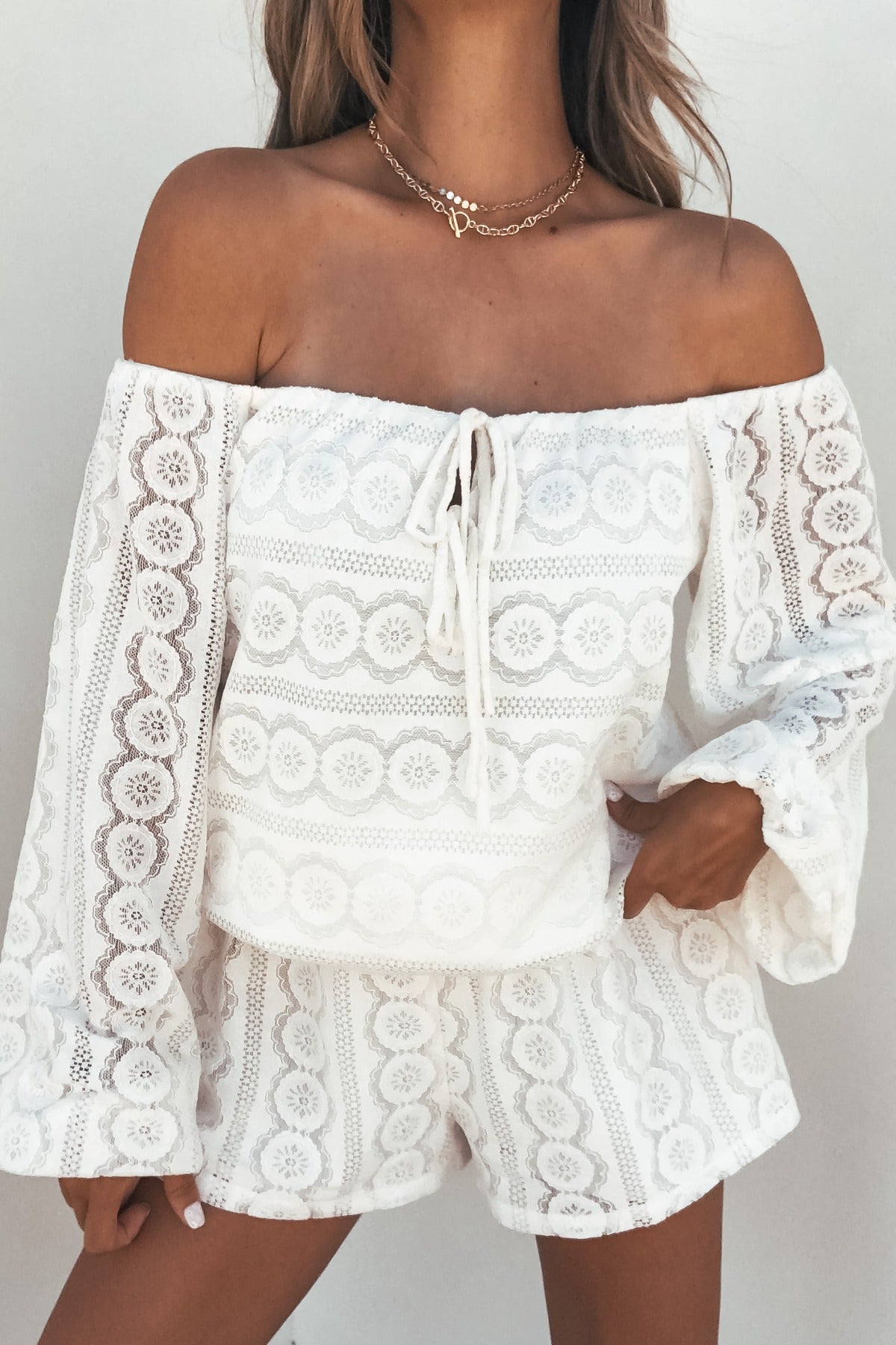 Woman wears the Elara Lace Off‑The‑Shoulder Romper in cream, styled with layered necklaces.