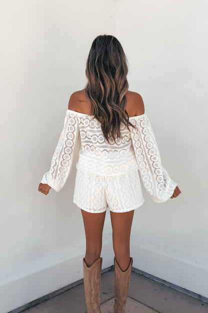 Woman in the cream Elara Lace Off-The-Shoulder Romper and tan cowboy boots stands against a white wall, facing away from the camera.