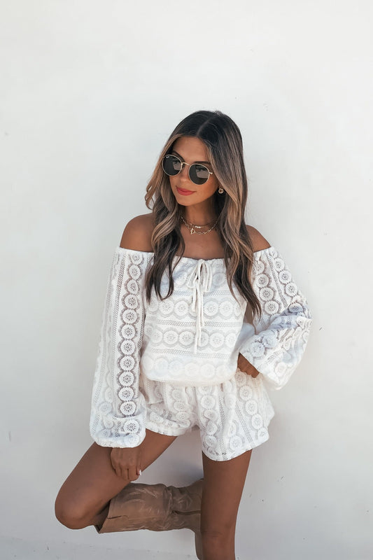 Woman in sunglasses and red lipstick wears the Elara Lace Off‑The‑Shoulder Romper – Cream with beige boots by a white wall.