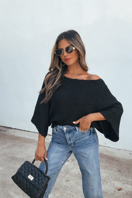 Woman in sunglasses wears the Elevated Ribbed Boatneck Sweater - Black with blue jeans and a quilted handbag.