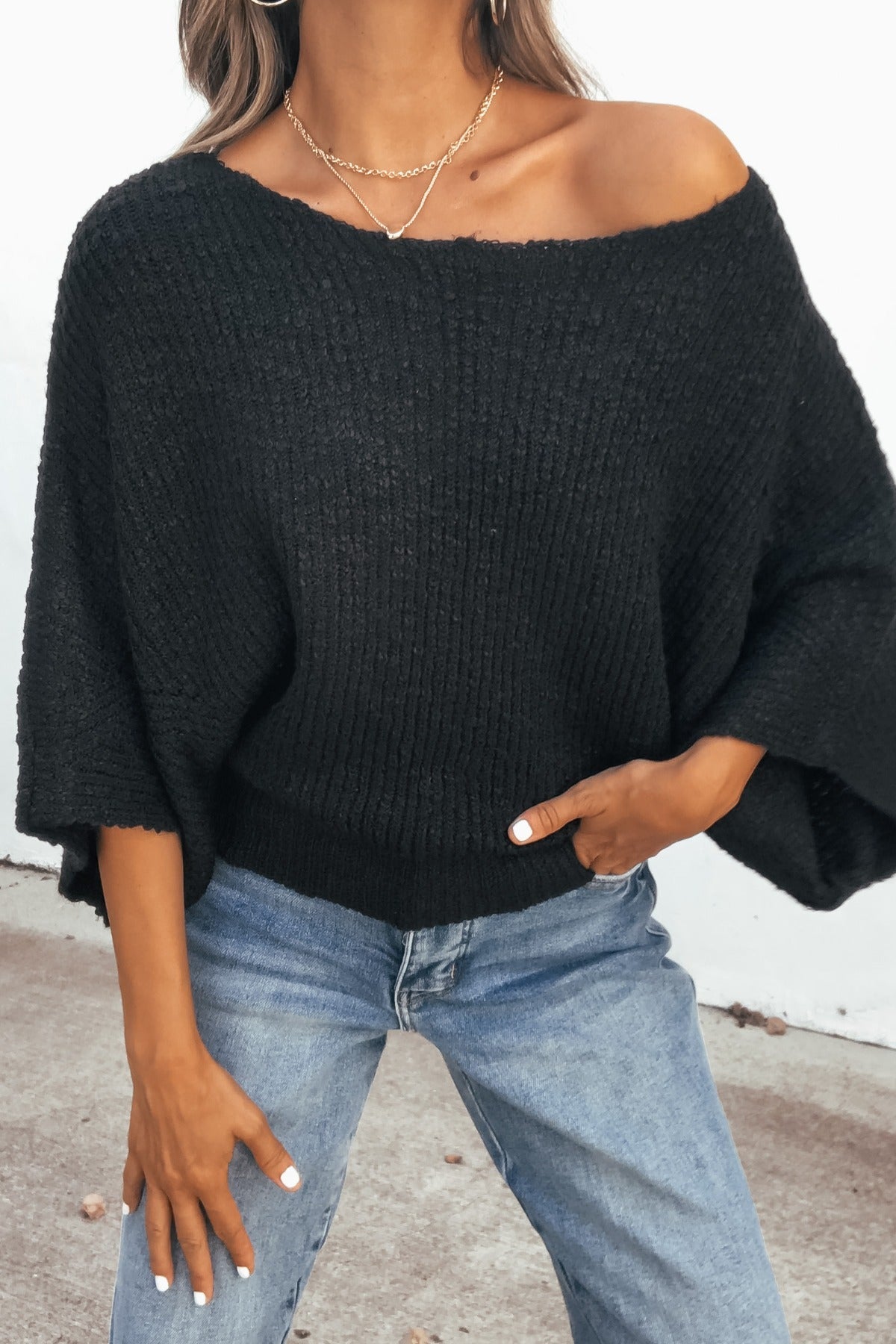 Woman wears the Elevated Ribbed Boatneck Sweater in black with blue jeans and necklaces, standing outdoors with a hand in her pocket.