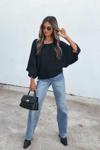 Woman in sunglasses and jeans poses confidently in an Elevated Ribbed Boatneck Sweater - Black, holding a quilted handbag by a white wall.