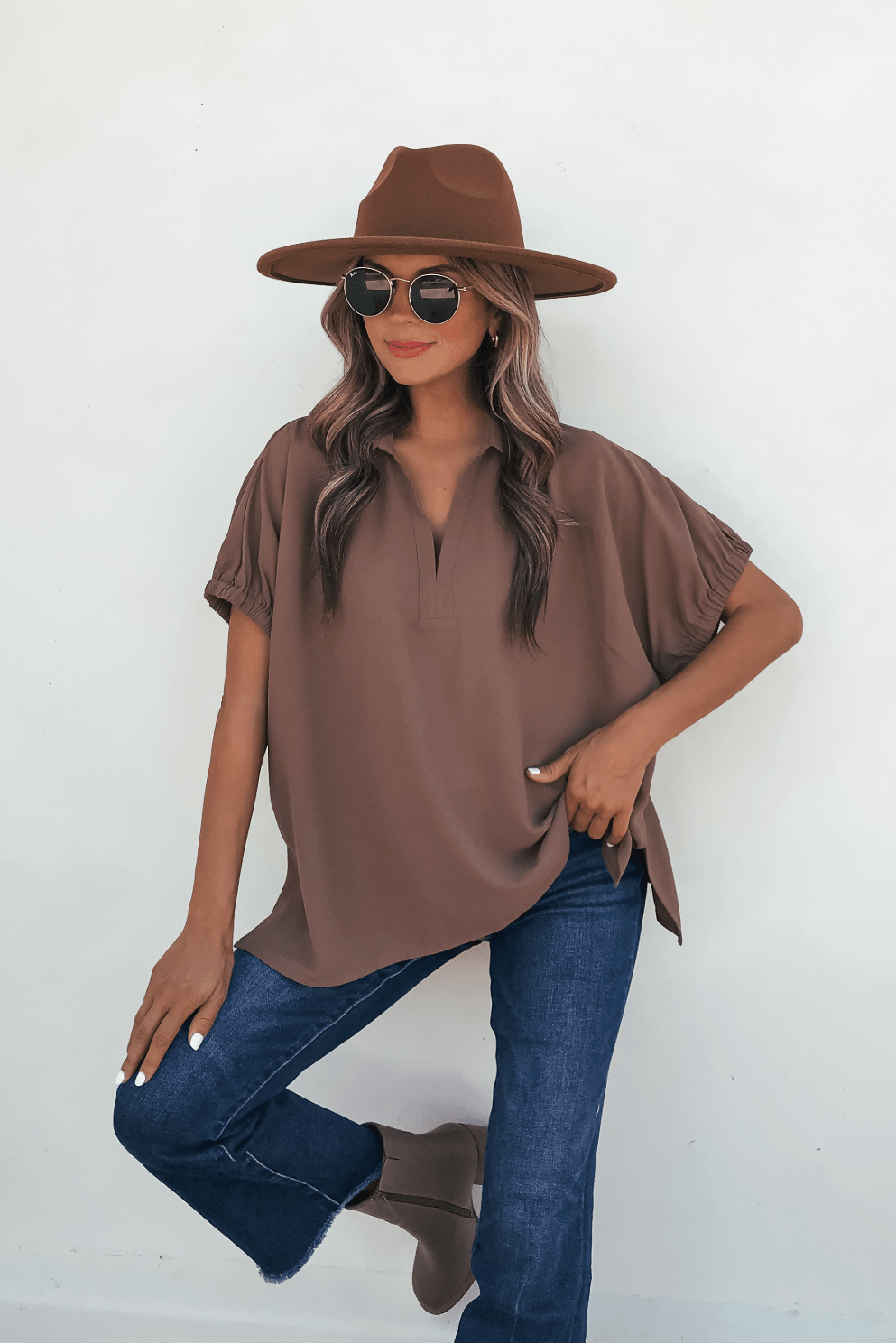 Woman in Ellis Camel Split Collared Top, wide-brim hat, and sunglasses poses by a white wall with one knee bent, hand on thigh.