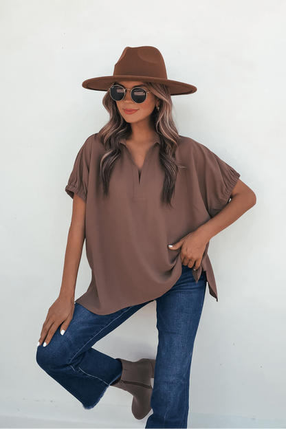 Woman in Ellis Camel Split Collared Top, wide-brim hat, and sunglasses poses by a white wall with one knee bent, hand on thigh.