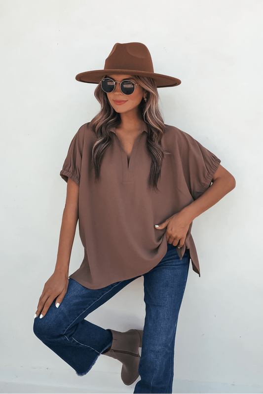 Woman in Ellis Camel Split Collared Top, wide-brim hat, and sunglasses poses by a white wall with one knee bent, hand on thigh.