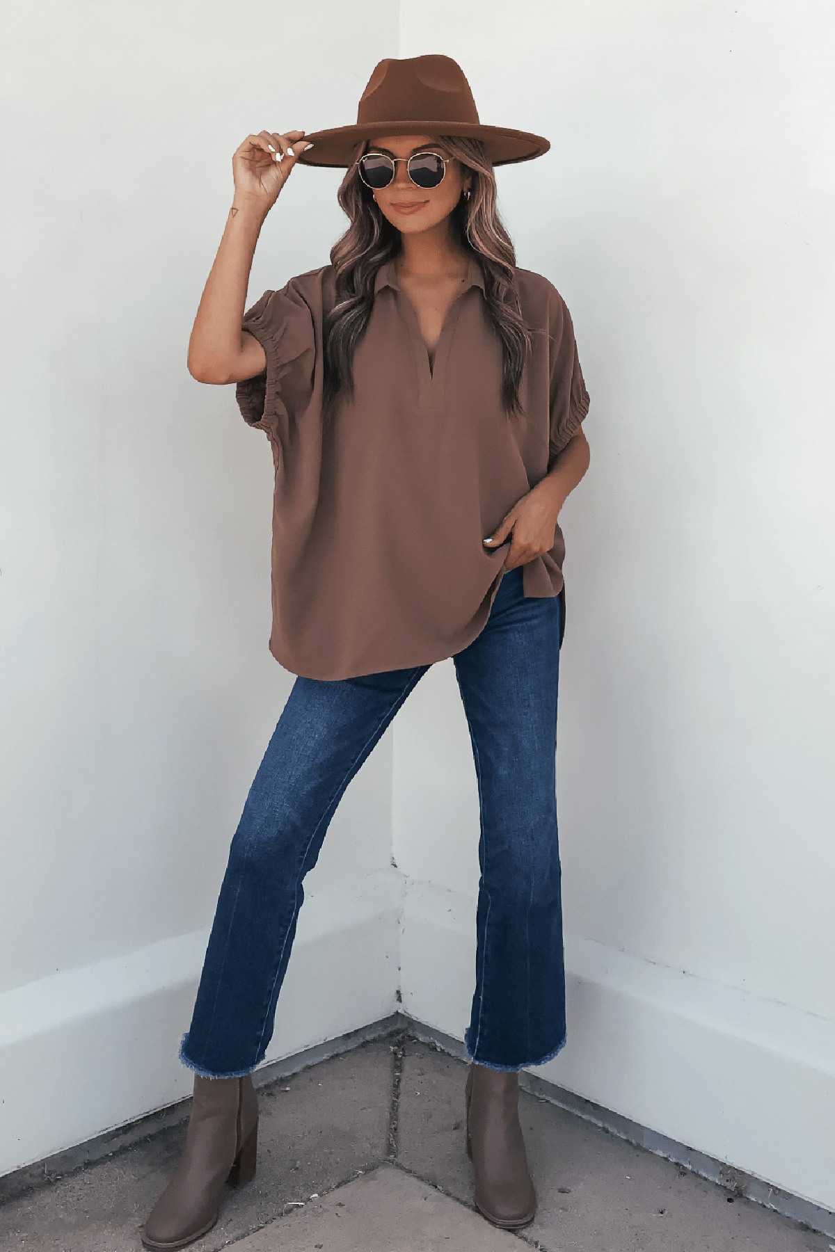 Woman wearing sunglasses, a brown hat, jeans, taupe boots, and the Ellis Camel Split Collared Top—an autumn essential by a white wall.