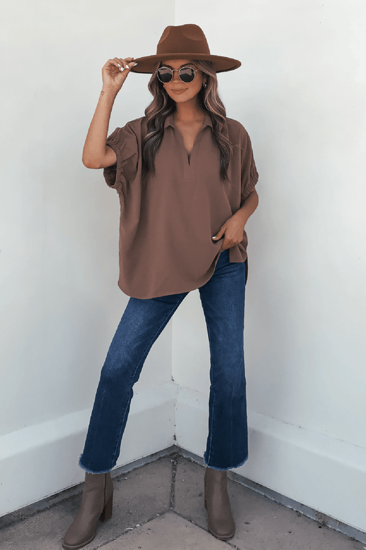 Woman in brown hat, sunglasses, Ellis Camel Split Collared Top, blue jeans, and brown boots poses by a white wall—autumn essential.