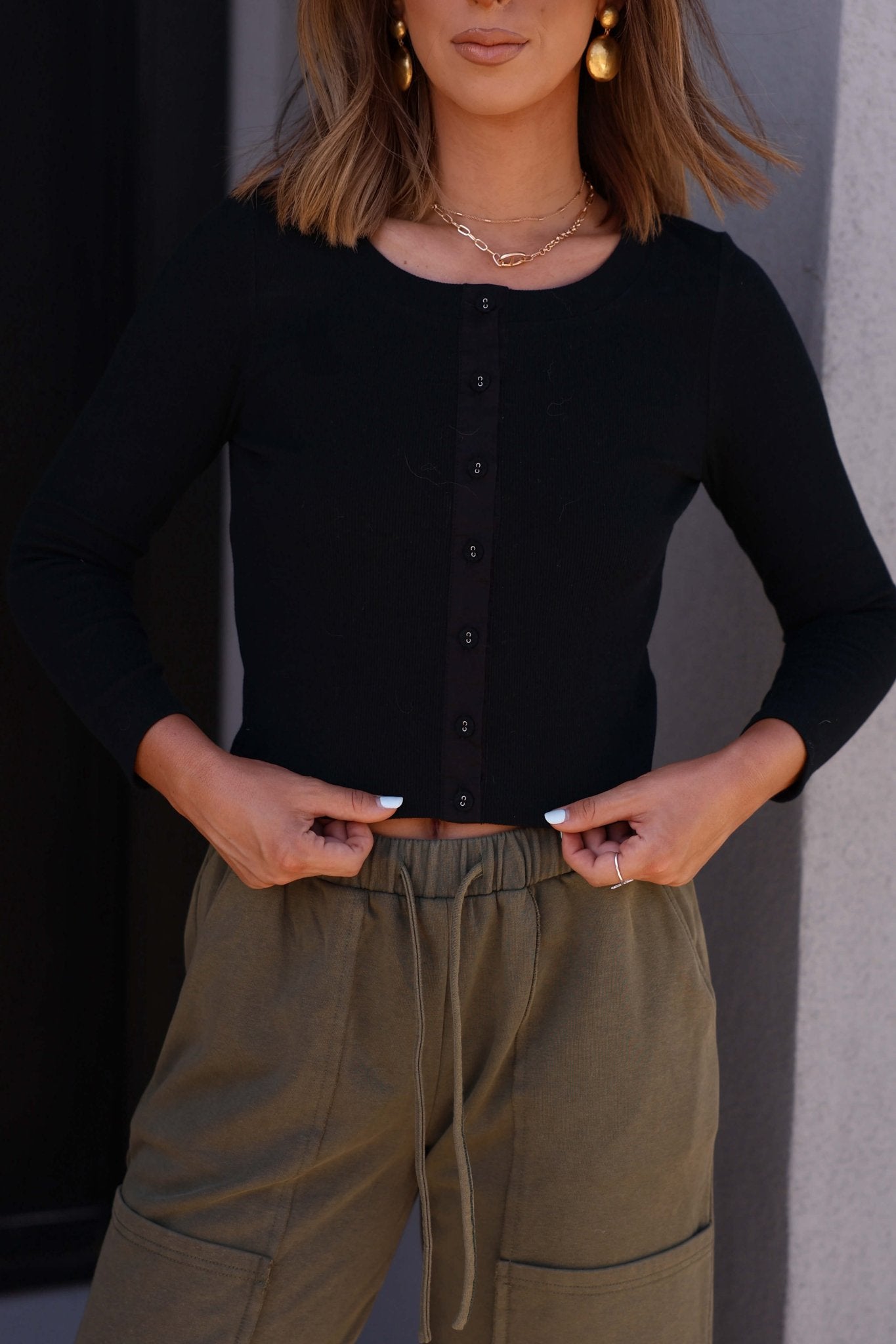 Woman wears the Eloise Black Ribbed Button Down Top, paired with olive green drawstring pants, against a light wall.