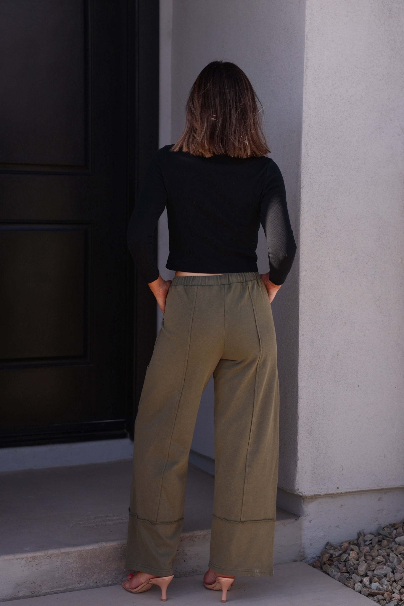 A person with shoulder-length hair wears the Eloise Black Ribbed Button Down Top, standing on a doorstep facing away.