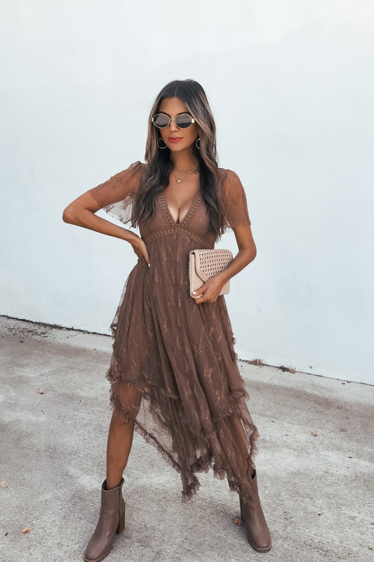 Woman in an Embellished Lace Midi Dress - Brown, with boots and clutch, sunglasses on, stands by a plain light wall.