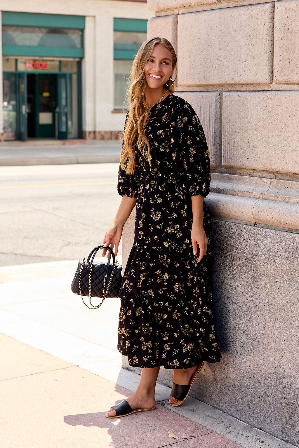 Woman in the Ember Floral Midi Dress - Black, sandals, and a black handbag smiles by a building.