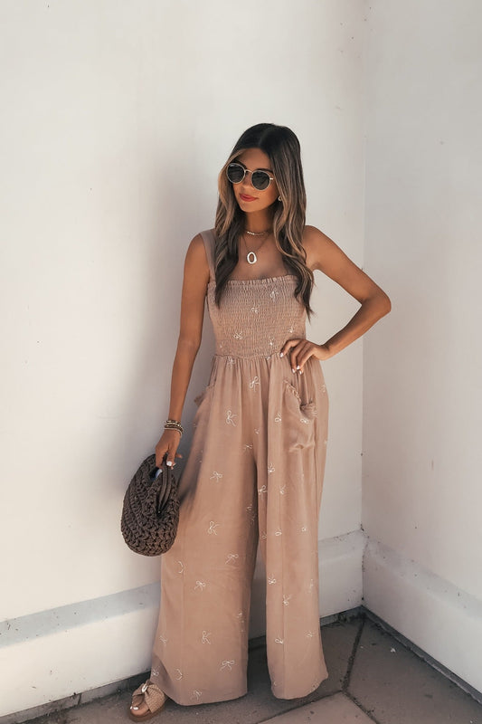 Woman in sunglasses, sandals, and a brown bag wears the Embroidered Smocked Linen Jumpsuit - Latte against an off-white wall.