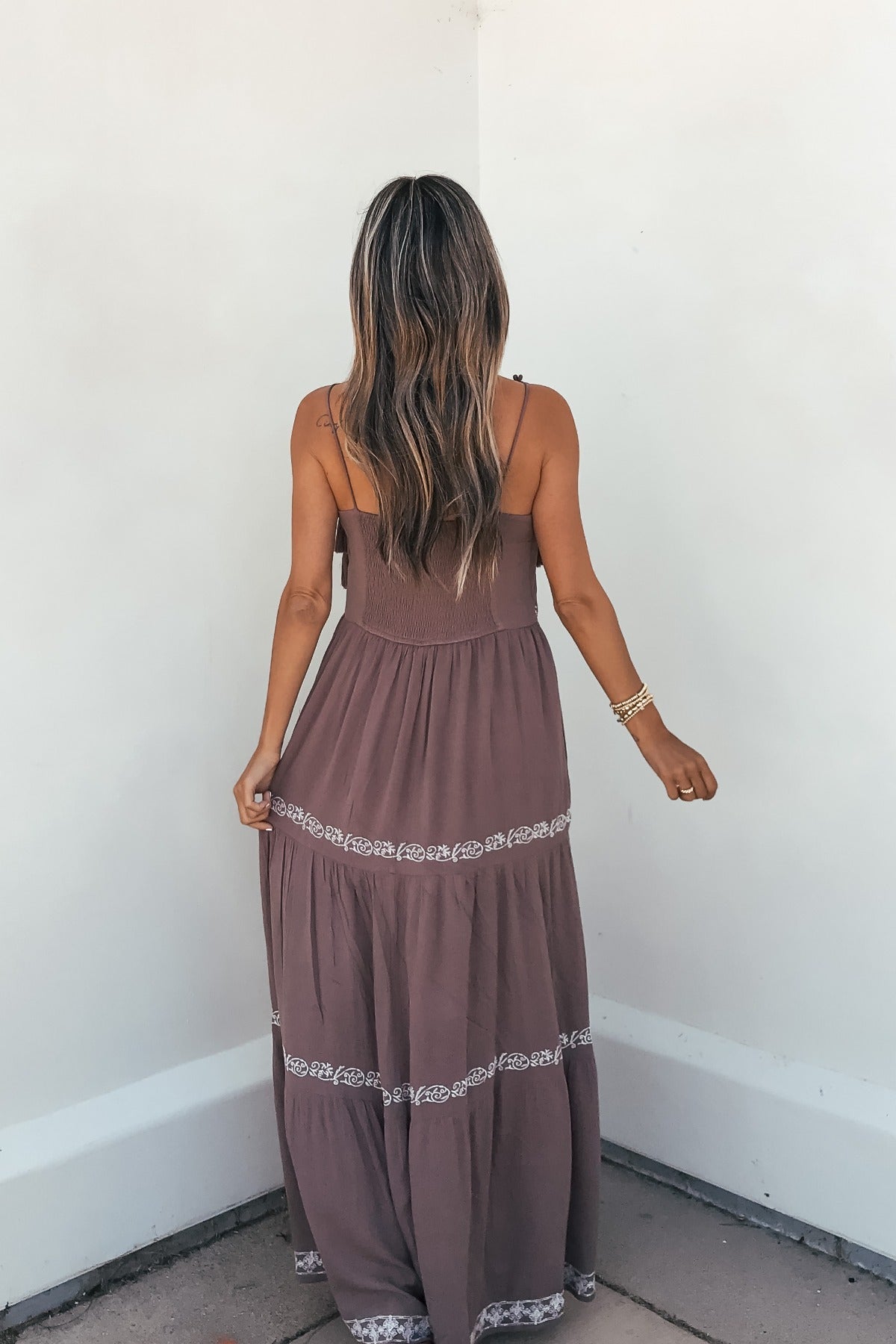 Woman with long, wavy hair wears an Embroidered Tiered Maxi Sun Dress - Mocha, standing against a white wall, facing away.