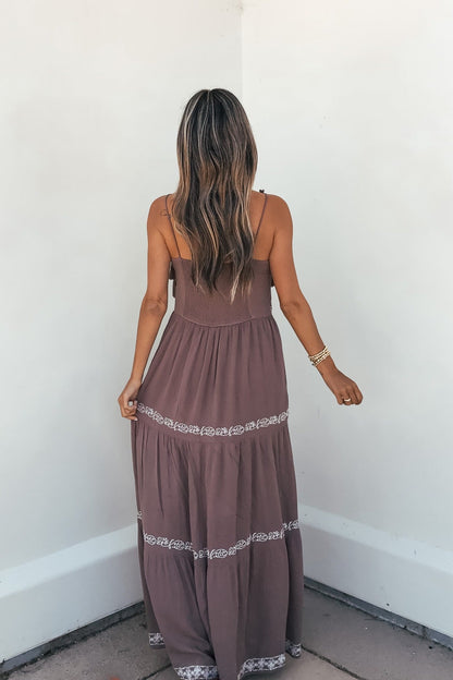Woman with long, wavy hair wears an Embroidered Tiered Maxi Sun Dress - Mocha, standing against a white wall, facing away.