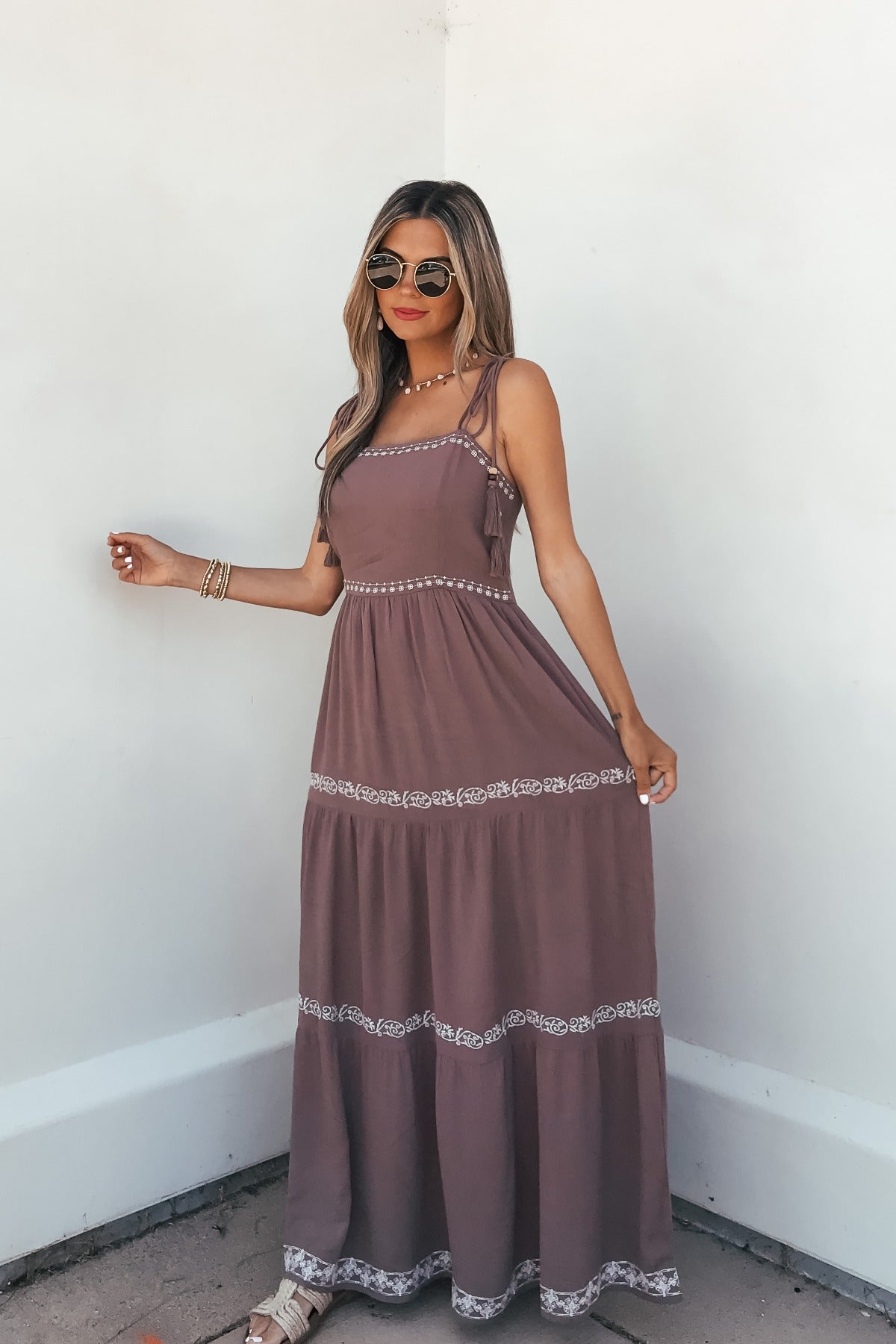 Woman in sunglasses poses against a white wall, wearing the Embroidered Tiered Maxi Sun Dress - Mocha and holding her summer dress.