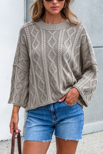 Woman in Emory Cable Knit Sweater - Olive, denim shorts, holding a brown bag by a concrete wall.