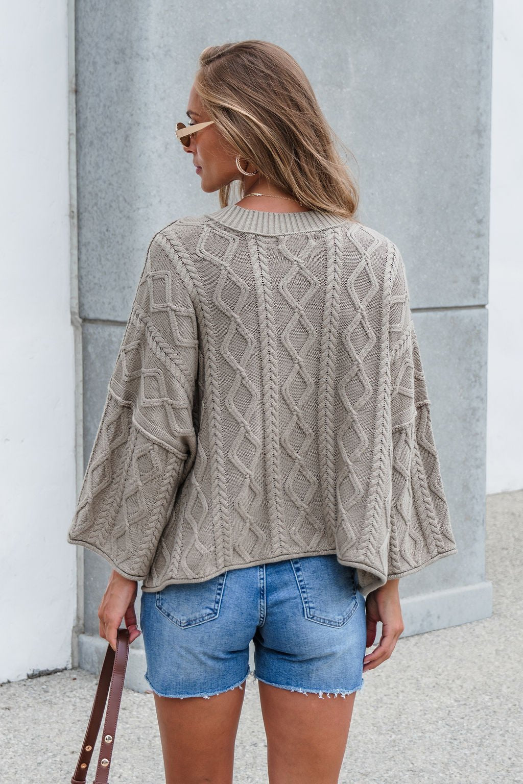 Woman in the Emory Cable Knit Sweater - Olive and denim shorts stands outside, facing away, holding a brown strap.