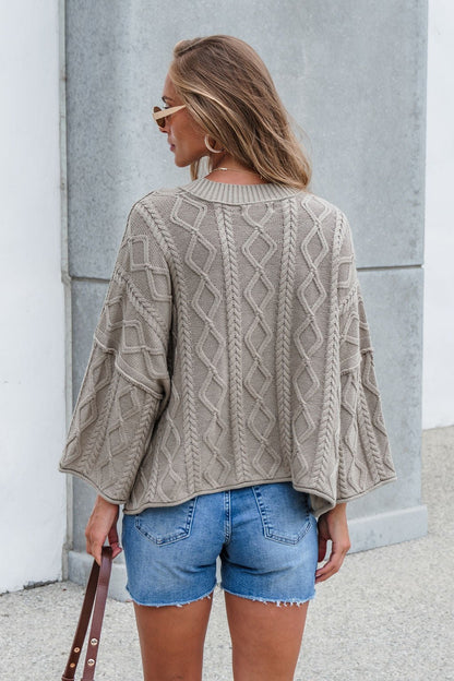 Woman in the Emory Cable Knit Sweater - Olive and denim shorts stands outside, facing away, holding a brown strap.