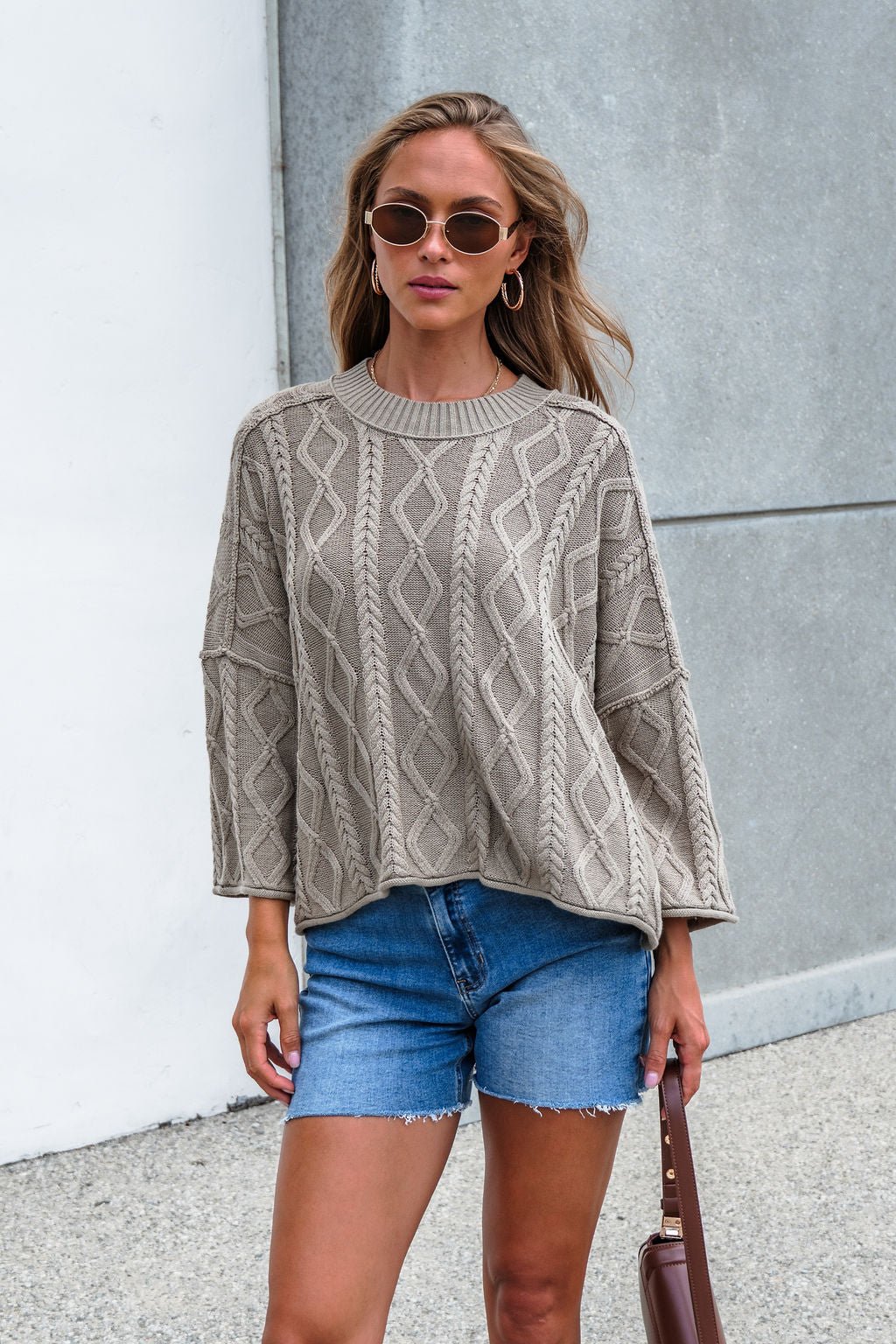 Woman in Emory Cable Knit Sweater - Olive and blue shorts stands outside against a white and gray wall.