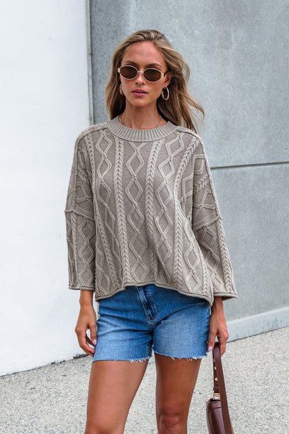 Woman in Emory Cable Knit Sweater - Olive and blue shorts stands outside against a white and gray wall.