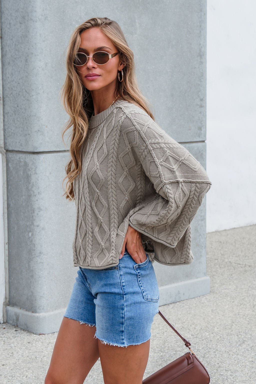 Woman in sunglasses wears the Emory Cable Knit Sweater in olive with denim shorts and a brown purse outdoors.