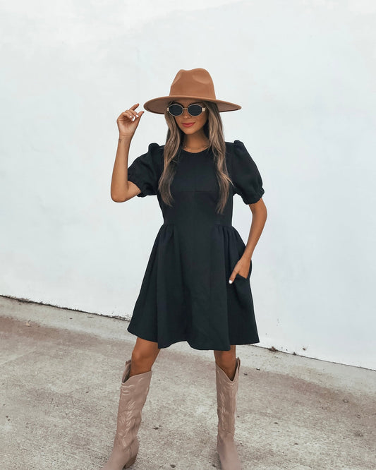 A person wearing a Puff Sleeve Mini Dress - Black, a tan fedora, sunglasses, and light-colored western boots.