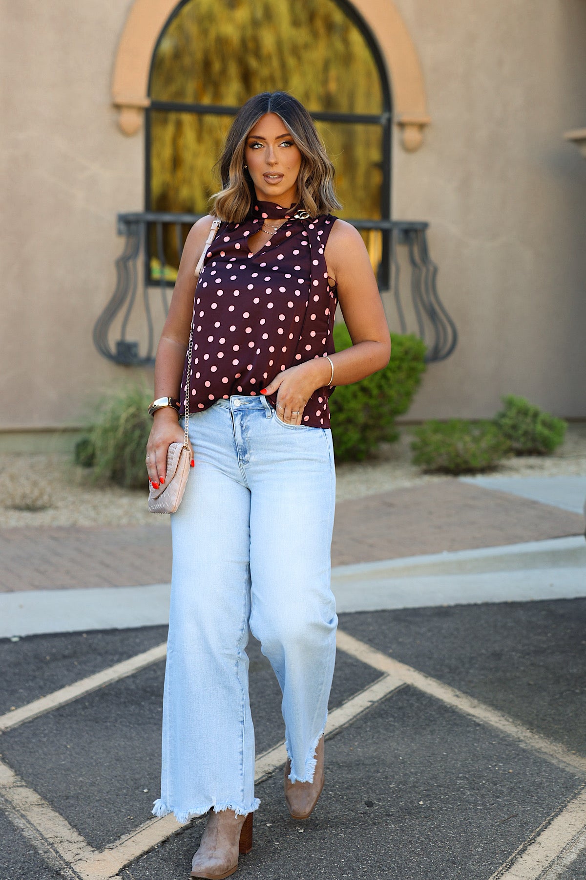 Woman wearing an ENTRO T33113 wine polka dot tank walks outdoors with light blue jeans, tan boots, and a pink clutch.