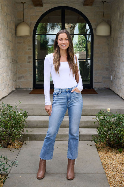 A woman smiles on stone steps outside, wearing the Essential Basic White Square Neck Top with blue jeans and brown boots.