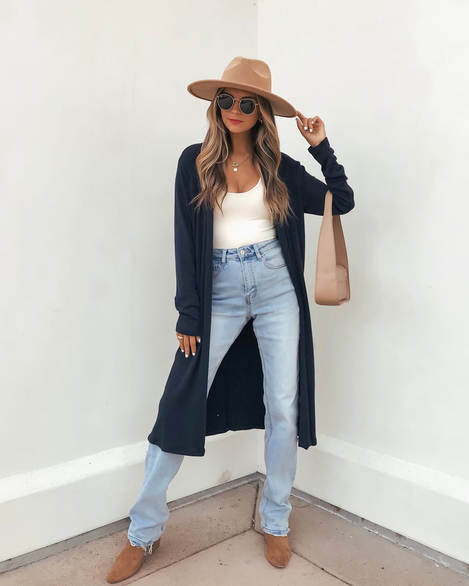Woman wearing the Essential Navy Duster Cardigan - FINAL SALE, tan hat, sunglasses, and light blue jeans poses by a white wall.
