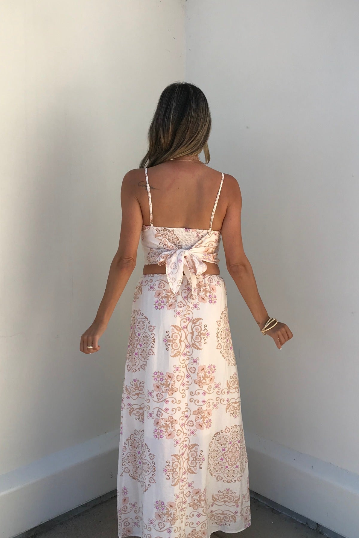 Woman in an Evelyn Cream Print Maxi Skirt, standing against a plain white wall, facing away.