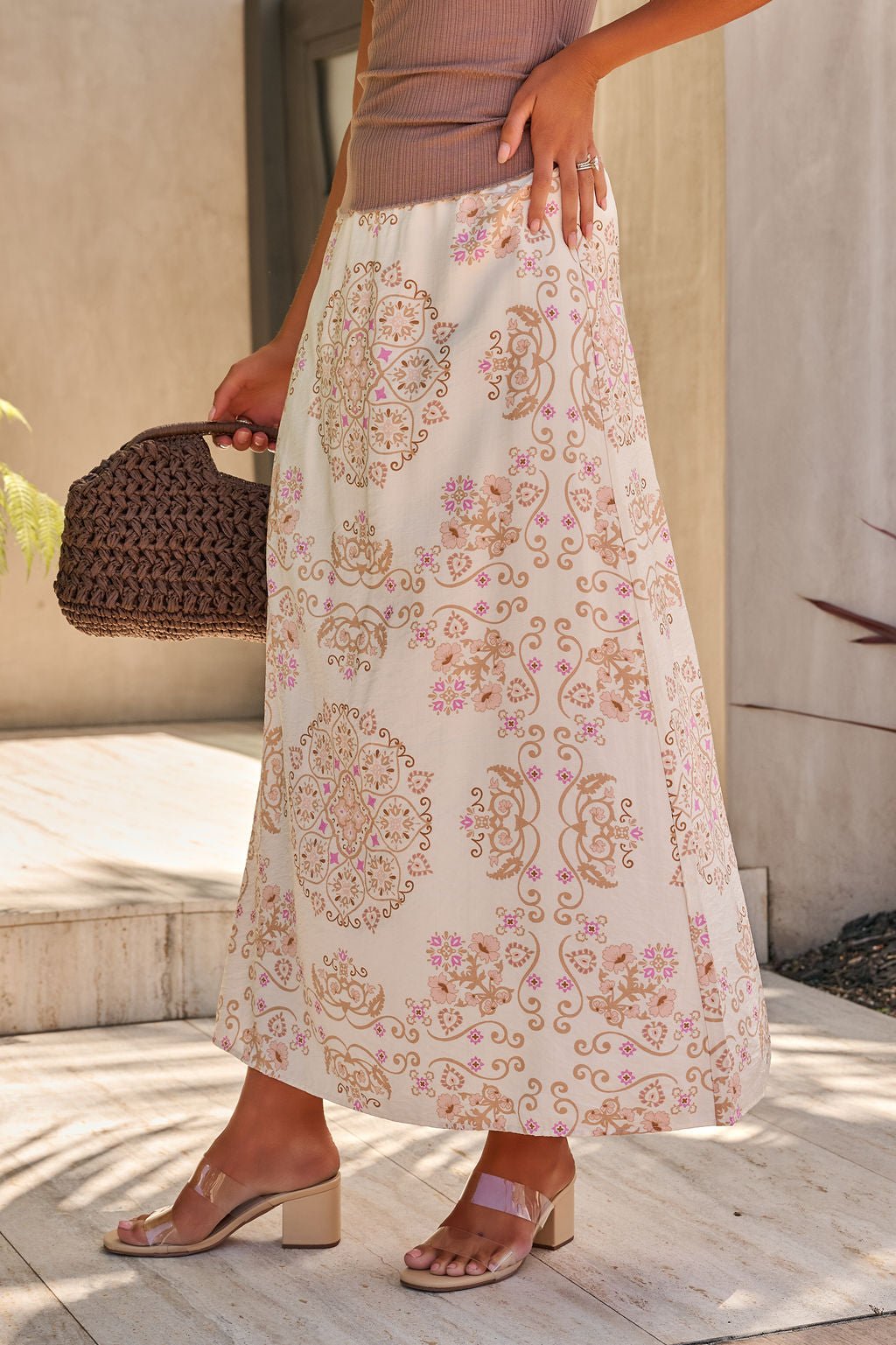 A woman outdoors in the Evelyn Cream Print Maxi Skirt with an elastic waistband, a woven handbag, and block-heeled sandals.