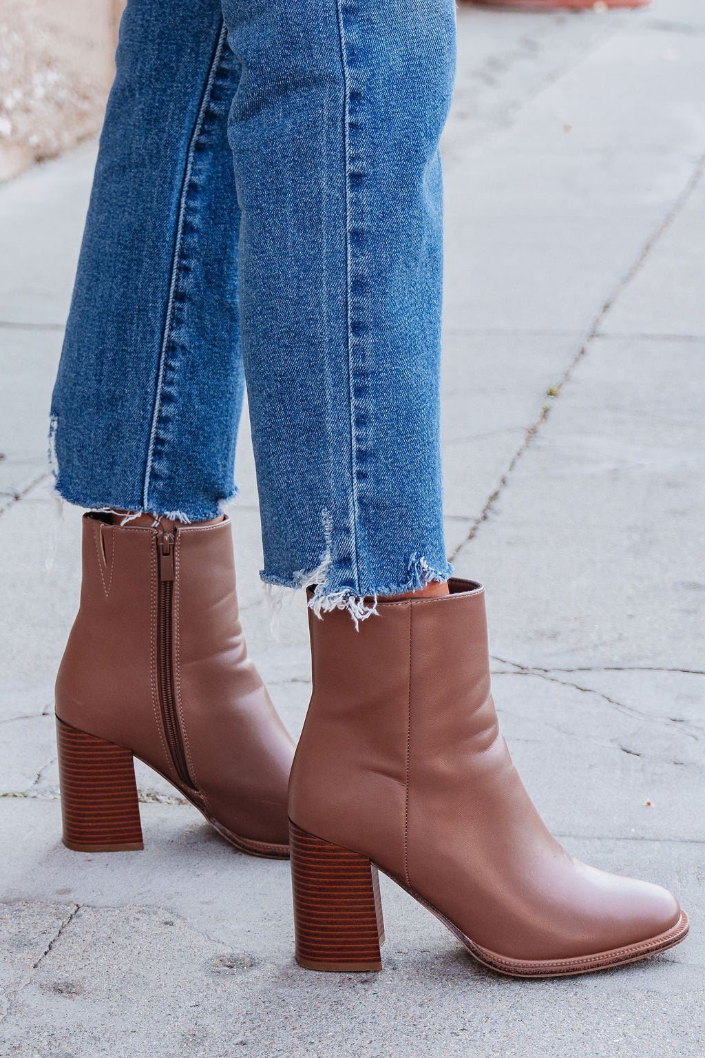 Person wearing frayed hem blue jeans and Evelyn Taupe Leather Heeled Ankle Booties, standing on a sidewalk.