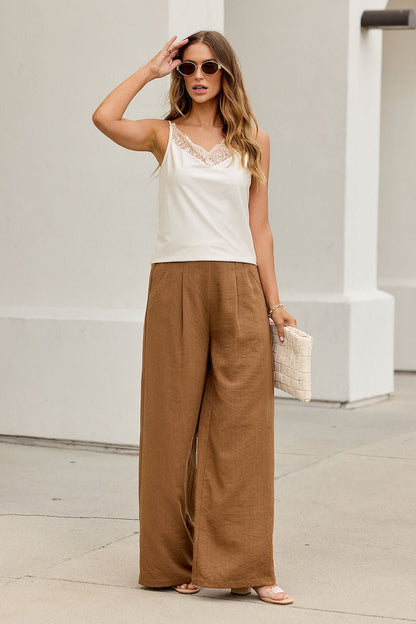 Woman in sunglasses wears the Evening Lace Satin Tank Top - Champagne with wide brown pants, sandals, and a woven clutch outdoors.