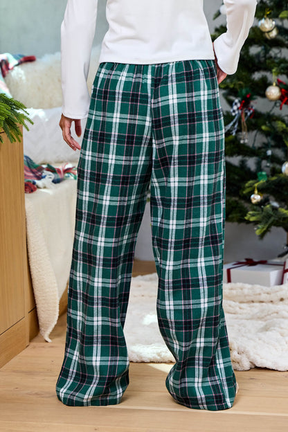 Wearing Evergreen Haven Plaid Lounge Pants, a person stands indoors by a festive Christmas tree and wrapped gifts.