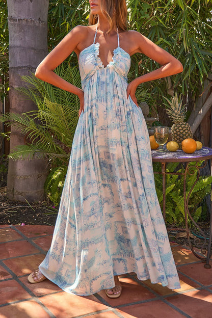 A woman in the Everlasting Print Maxi Dress - Blue with a halter neckline stands on a patio amid tropical plants, pineapple & oranges.