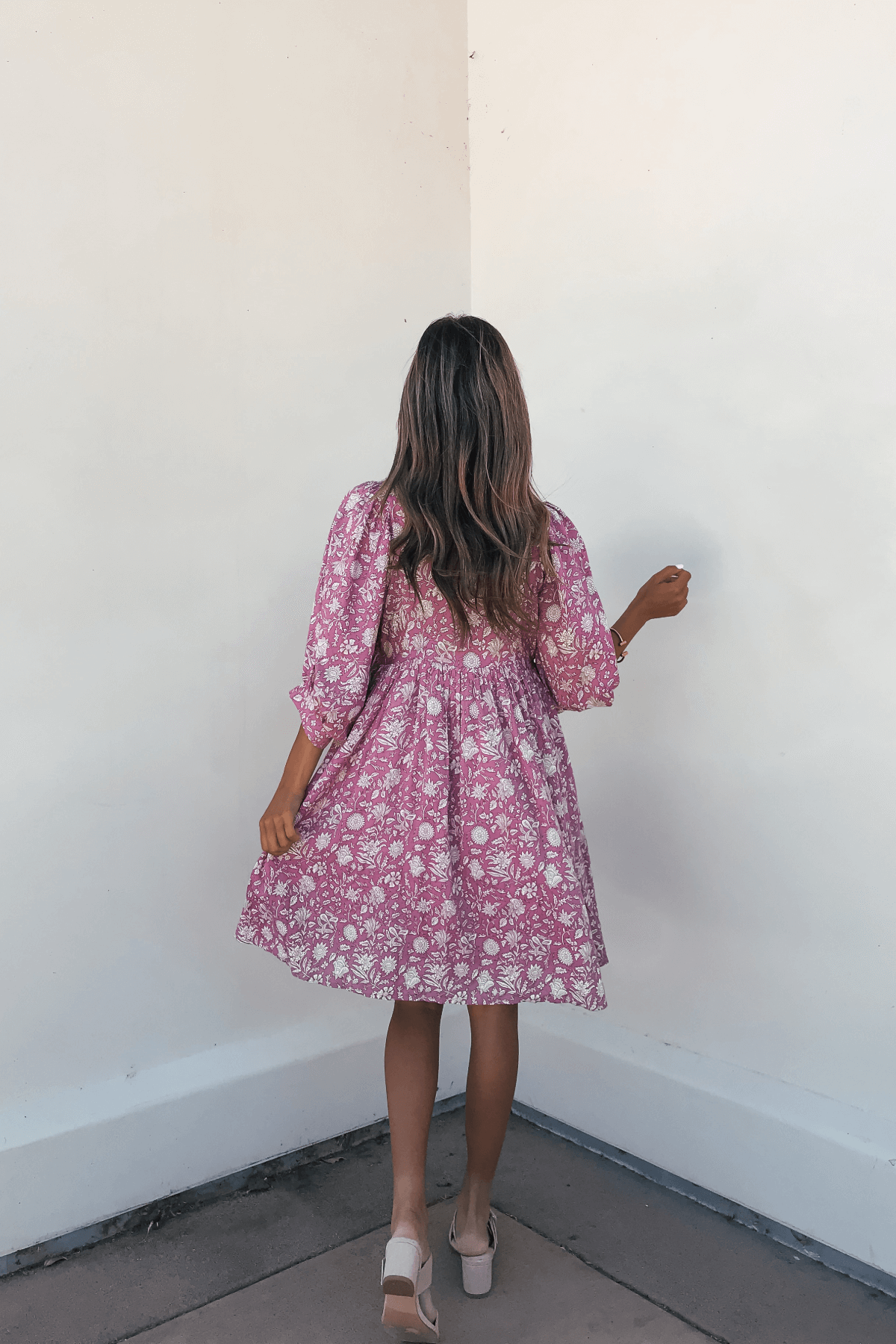 A woman in the Everly Pink Floral Print Mini Dress with flowy half sleeves stands facing a white wall, her back to the camera.