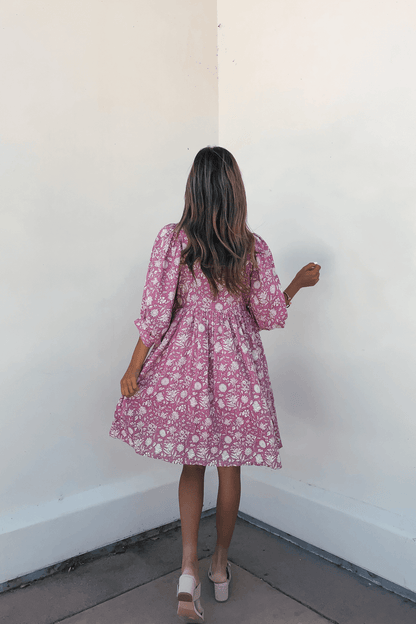 A woman in the Everly Pink Floral Print Mini Dress with flowy half sleeves stands facing a white wall, her back to the camera.