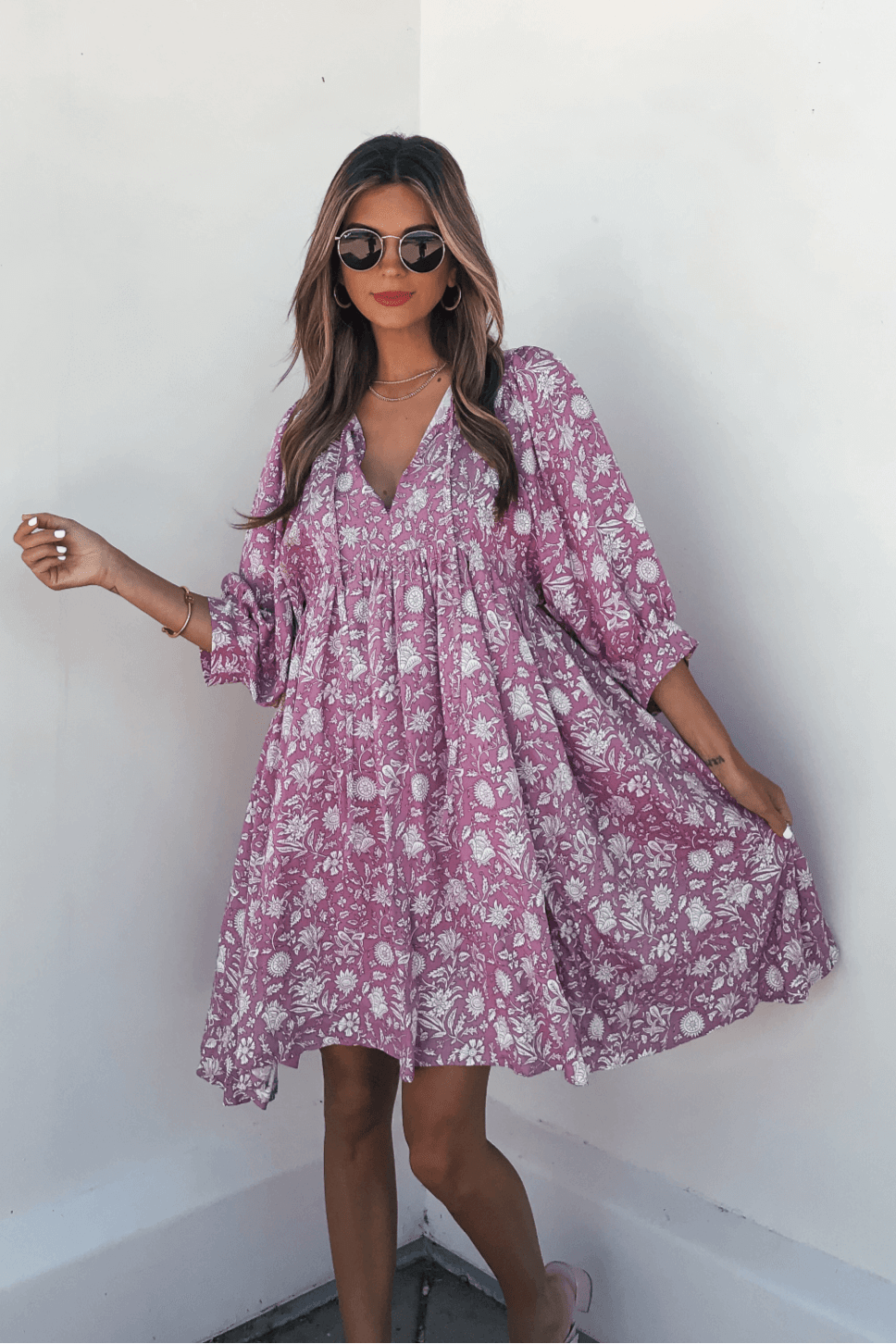 Woman wears the Everly Pink Floral Print Mini Dress with flowy sleeves and smiles as she poses against a white wall.