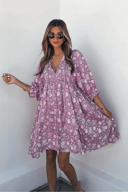 Woman wears the Everly Pink Floral Print Mini Dress with flowy sleeves and smiles as she poses against a white wall.
