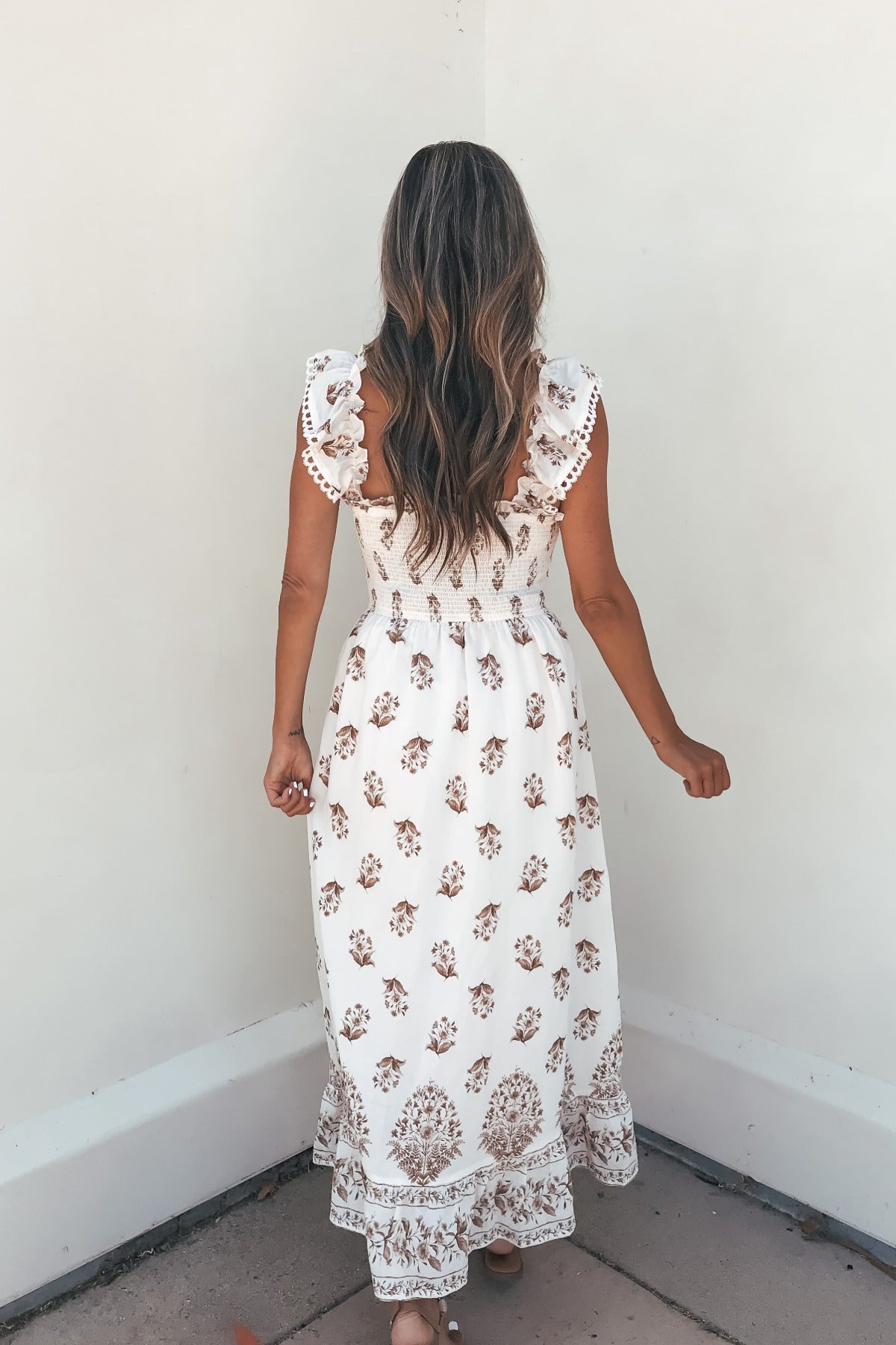 Woman with long wavy hair wears the Everly Smocked Print Midi Dress - Ivory, standing with her back to a white wall.