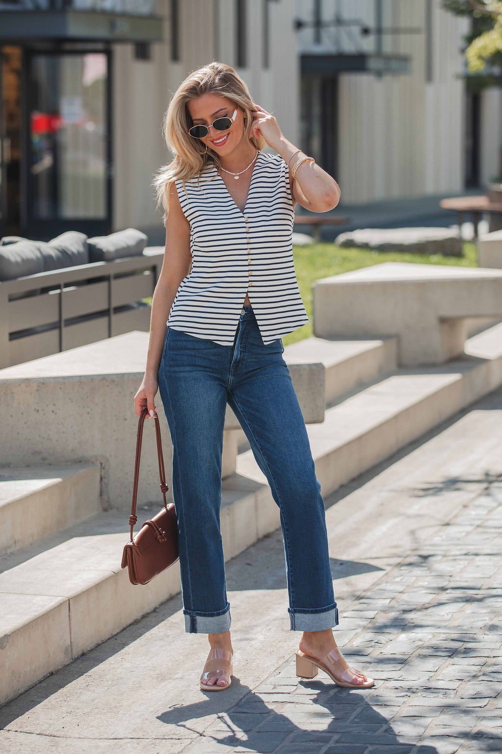 A woman in sunglasses wears a Fairview Stripe Buttoned Top - Navy, blue jeans, and sandals, carrying a brown handbag outdoors.