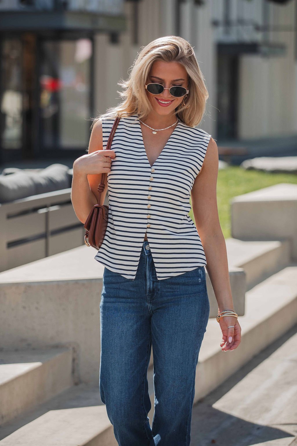 Woman in sunglasses wearing the Fairview Stripe Buttoned Top - Navy and blue jeans, walking outdoors on a sunny day.