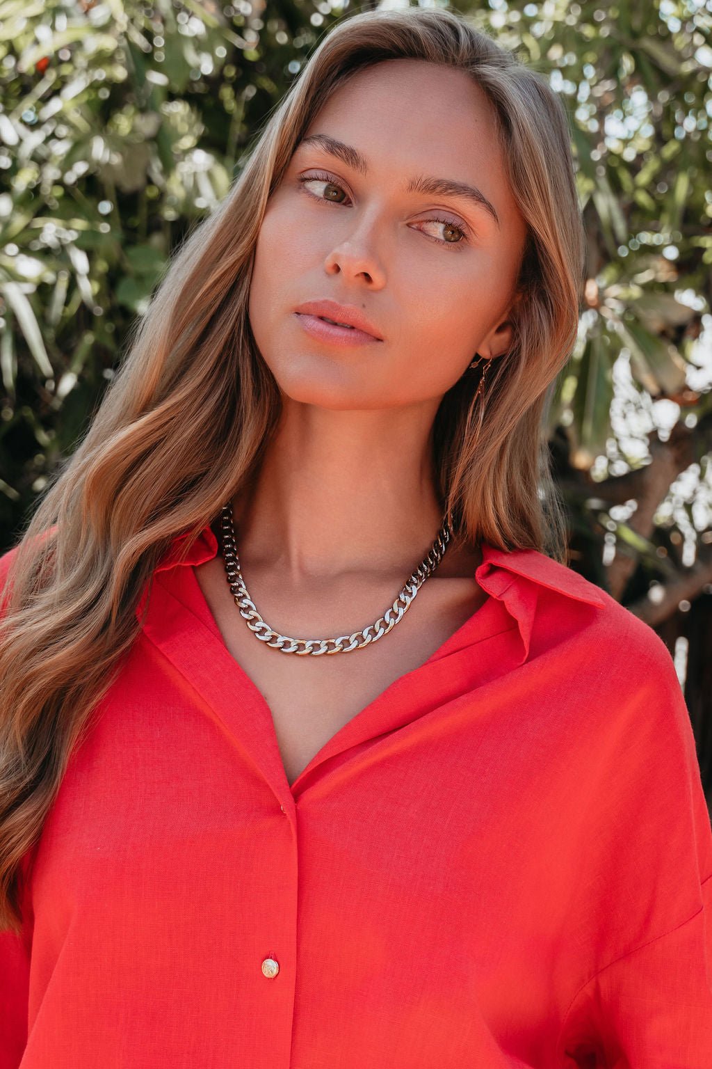 Woman with long, wavy hair wears a bright red shirt and the Farrah B Gold Plated Mixed Metal Chain Necklace outdoors by green foliage.