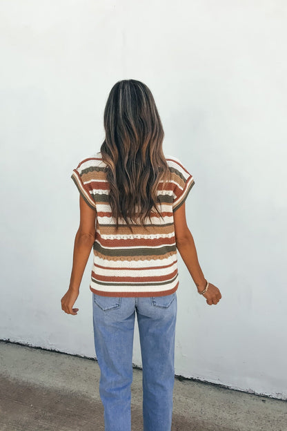 A woman with long wavy hair wears the Field Day Stripe Sweater Vest in olive and blue jeans, standing against a white wall, facing away.